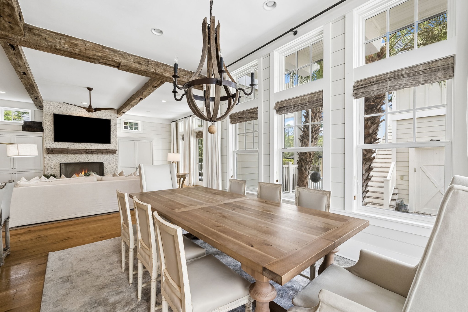 Gather around the rustic dining table beneath soaring wood beams and a statement chandelier, with the fireplace glowing nearby and palm trees swaying just beyond your windows.