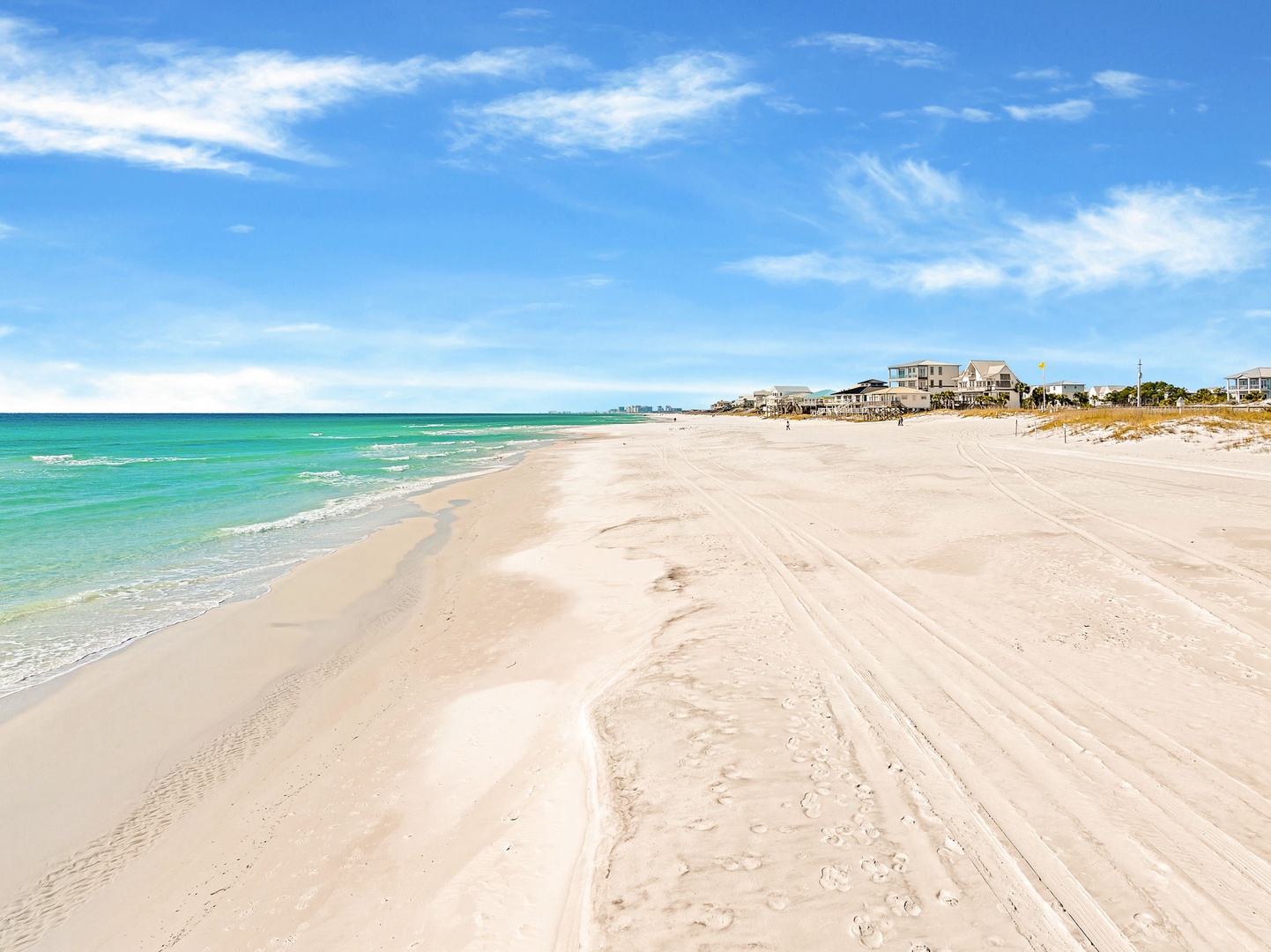 Pristine white sand beach stretches endlessly along turquoise waters, with coastal homes dotting the shoreline under brilliant blue skies.