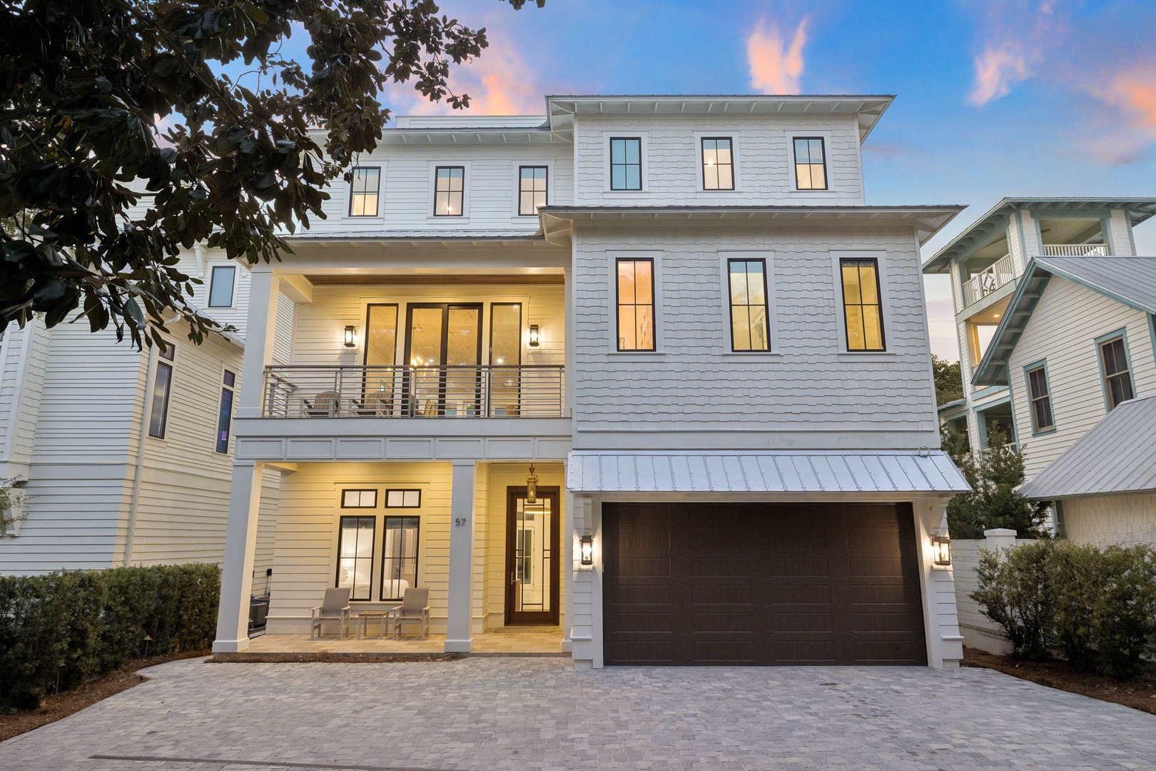 Modern three-story coastal home with elegant white siding, multiple balconies, and warm evening lighting in a charming neighborhood setting.