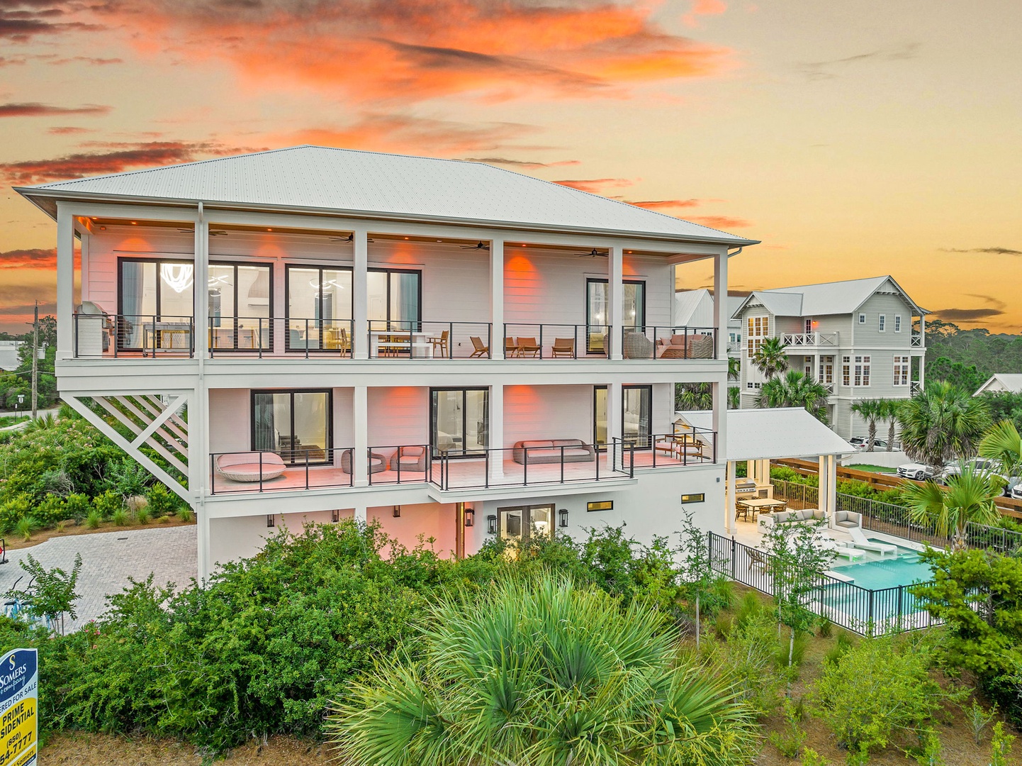 A stunning three-story coastal property features expansive balconies, tropical landscaping, and a private pool, set against a dramatic sunset sky in a peaceful neighborhood.