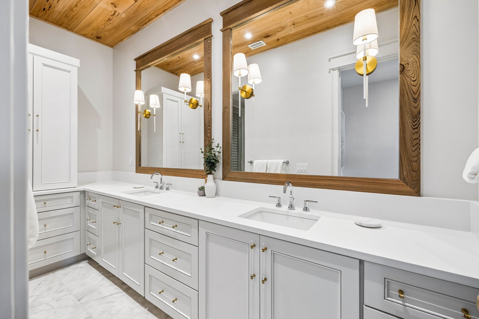 Your private bathroom features dual vanities with elegant white quartz counters, custom wood-framed mirrors reflecting the warm wood-beam ceiling, and stylish gold accent sconces adding a soft, welcoming glow.