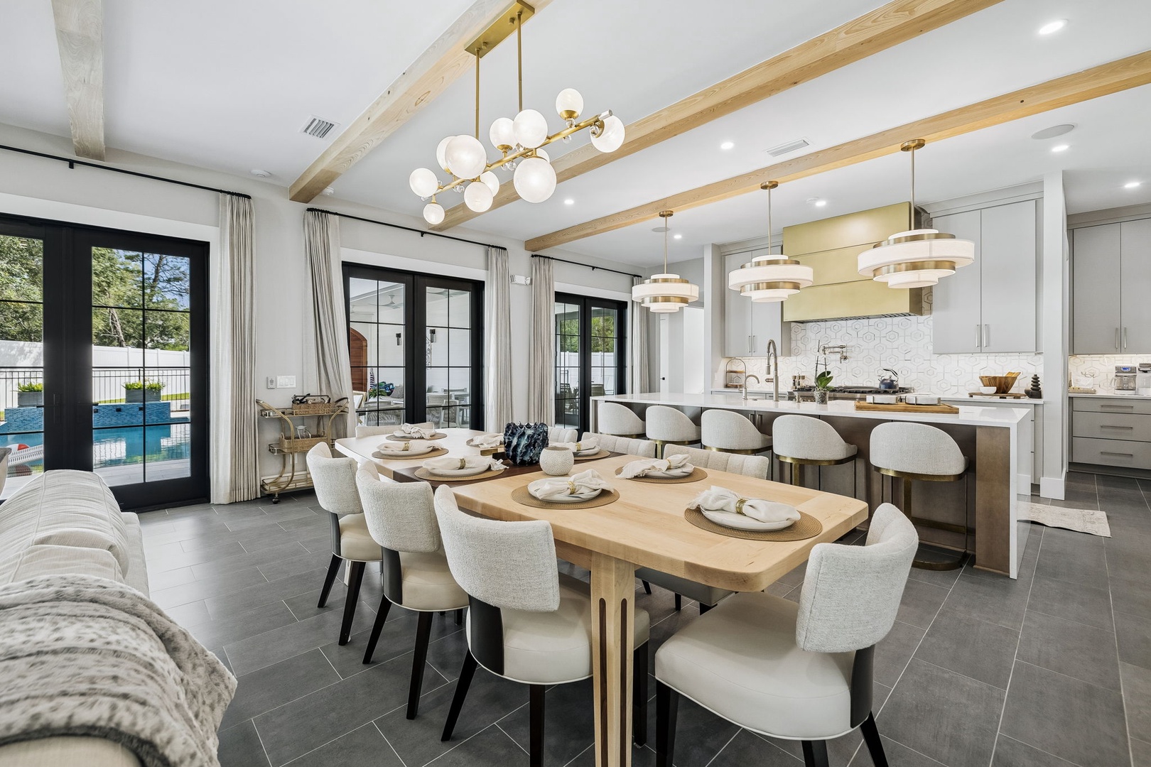 Stunning dining area with modern kitchen island and wood beam ceilings. Natural light floods through glass doors revealing the sparkling pool beyond, creating an elegant setting for memorable meals.