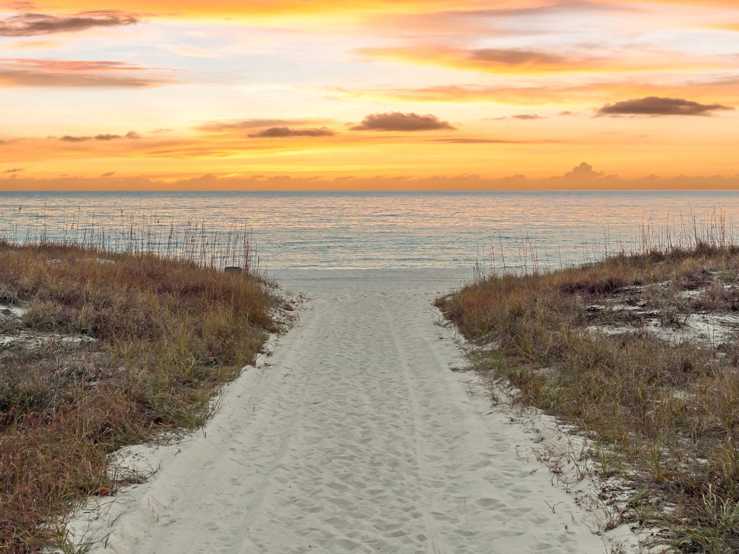 Stunning sunset paints the sky golden above pristine beach access, with sandy path leading to tranquil waters.