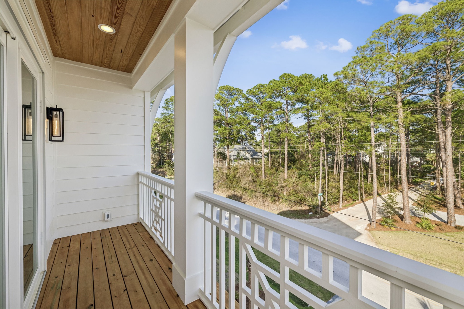 Covered balcony with wooden decking overlooking a peaceful wooded area with tall trees and blue sky.
