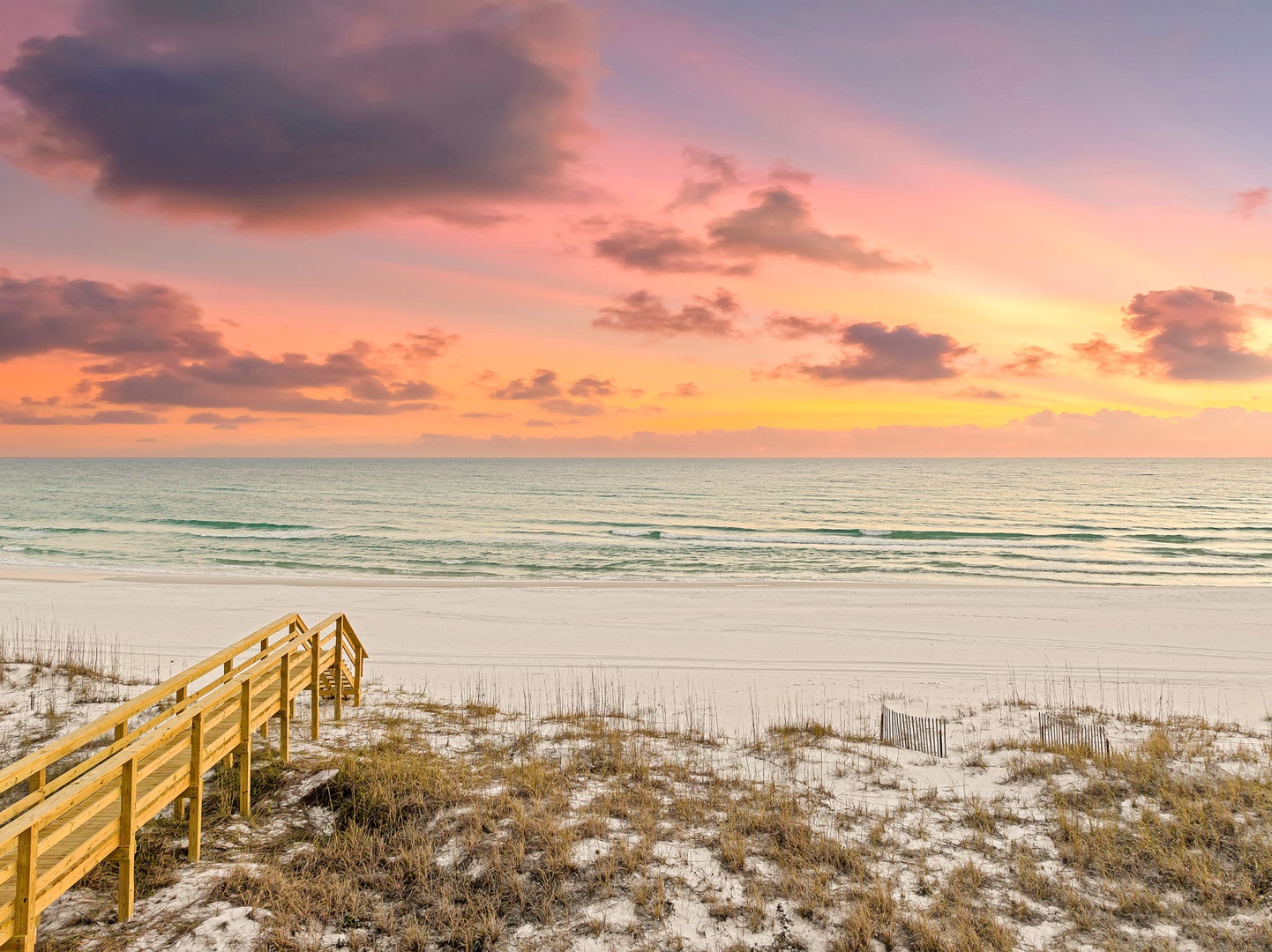 Stunning sunset paints the sky in brilliant hues over pristine white sand beach with wooden boardwalk leading to the shore.