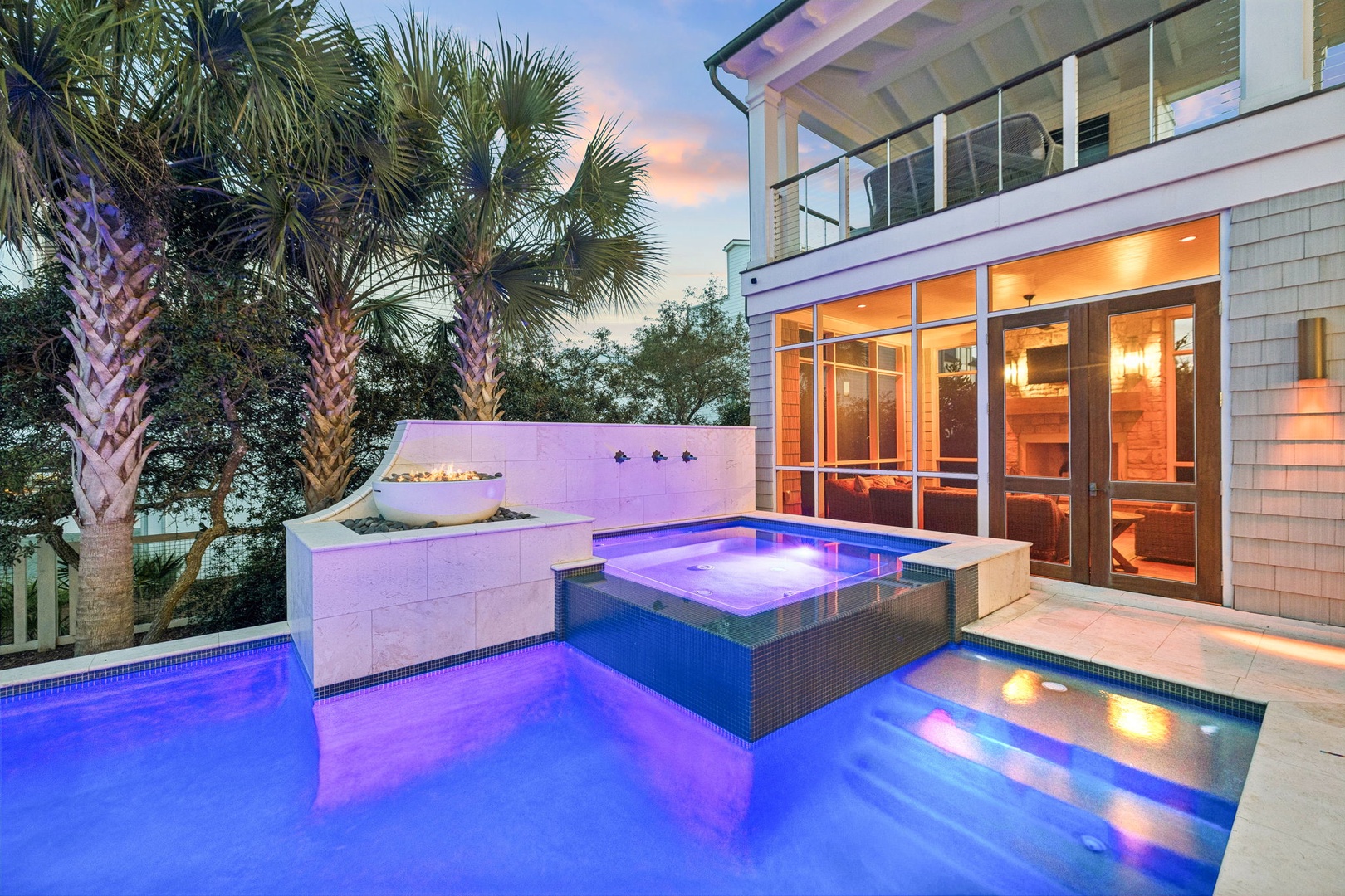 Modern coastal property featuring an elegant pool and spa area surrounded by tropical palms, with contemporary architecture and warm interior lighting visible through expansive glass doors at dusk.