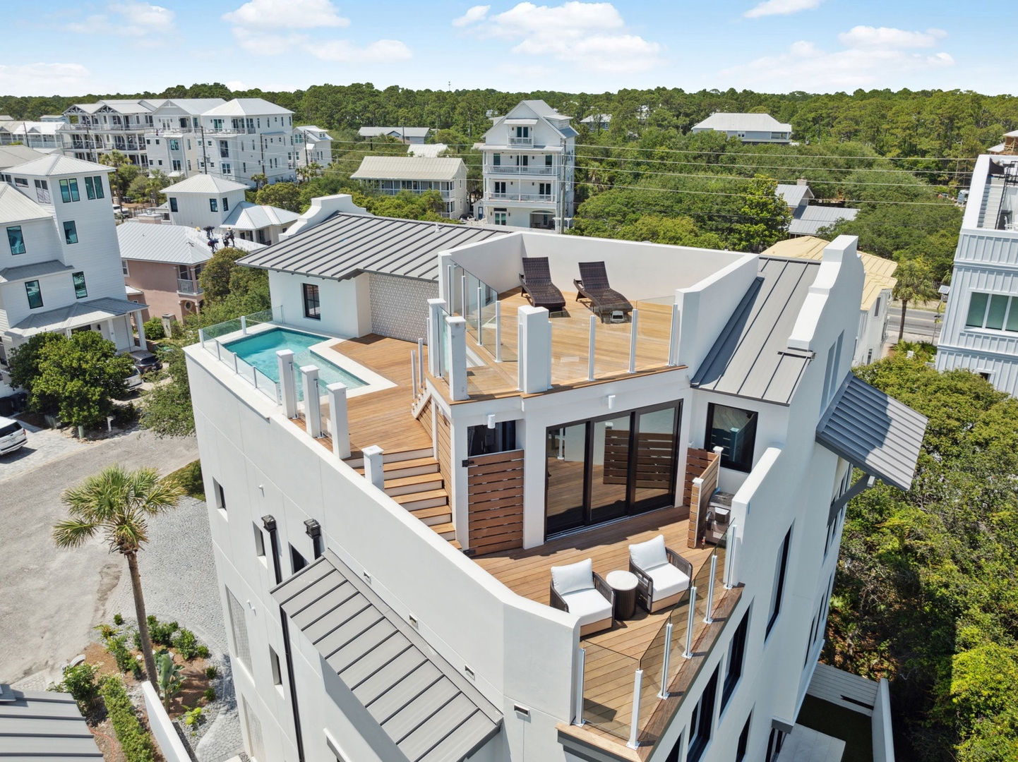 A modern beach home with expansive rooftop deck, pool, and sunbeds in a coastal neighborhood. The property features contemporary architecture with multiple levels, surrounded by lush greenery and nearby vacation rentals.