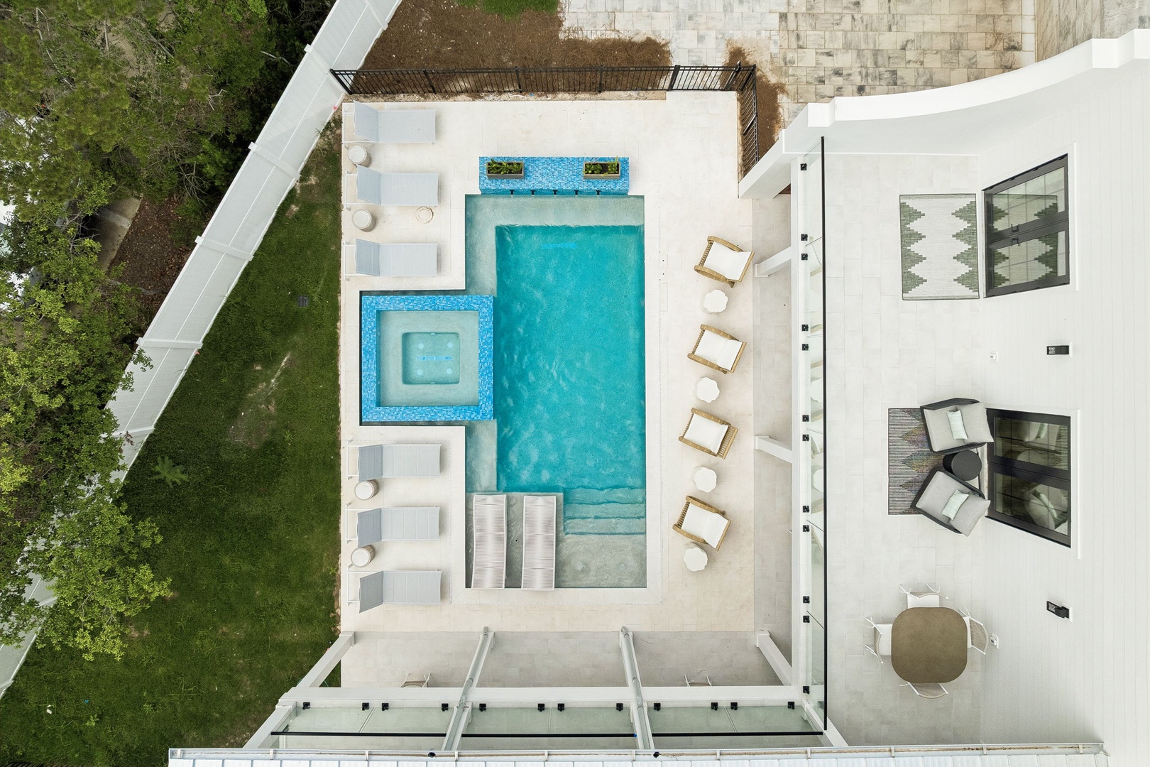 Aerial view of a modern vacation rental featuring a rectangular swimming pool with an adjacent square hot tub, surrounded by loungers and an outdoor dining terrace, nestled among lush greenery.