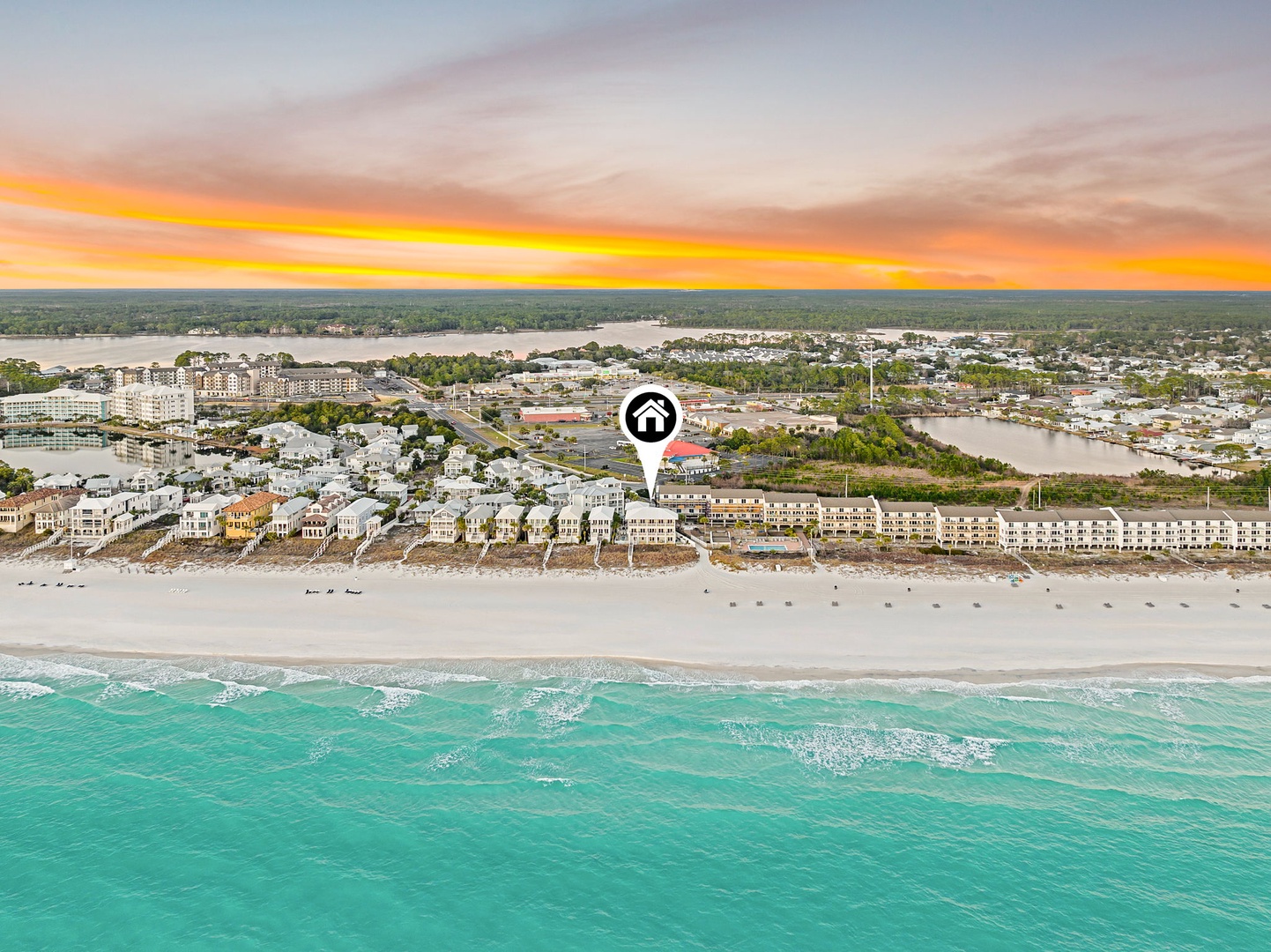 Aerial view of beachfront property located on pristine white sand shores with turquoise waters. Coastal community features nearby waterways and residential areas under a stunning sunset horizon.