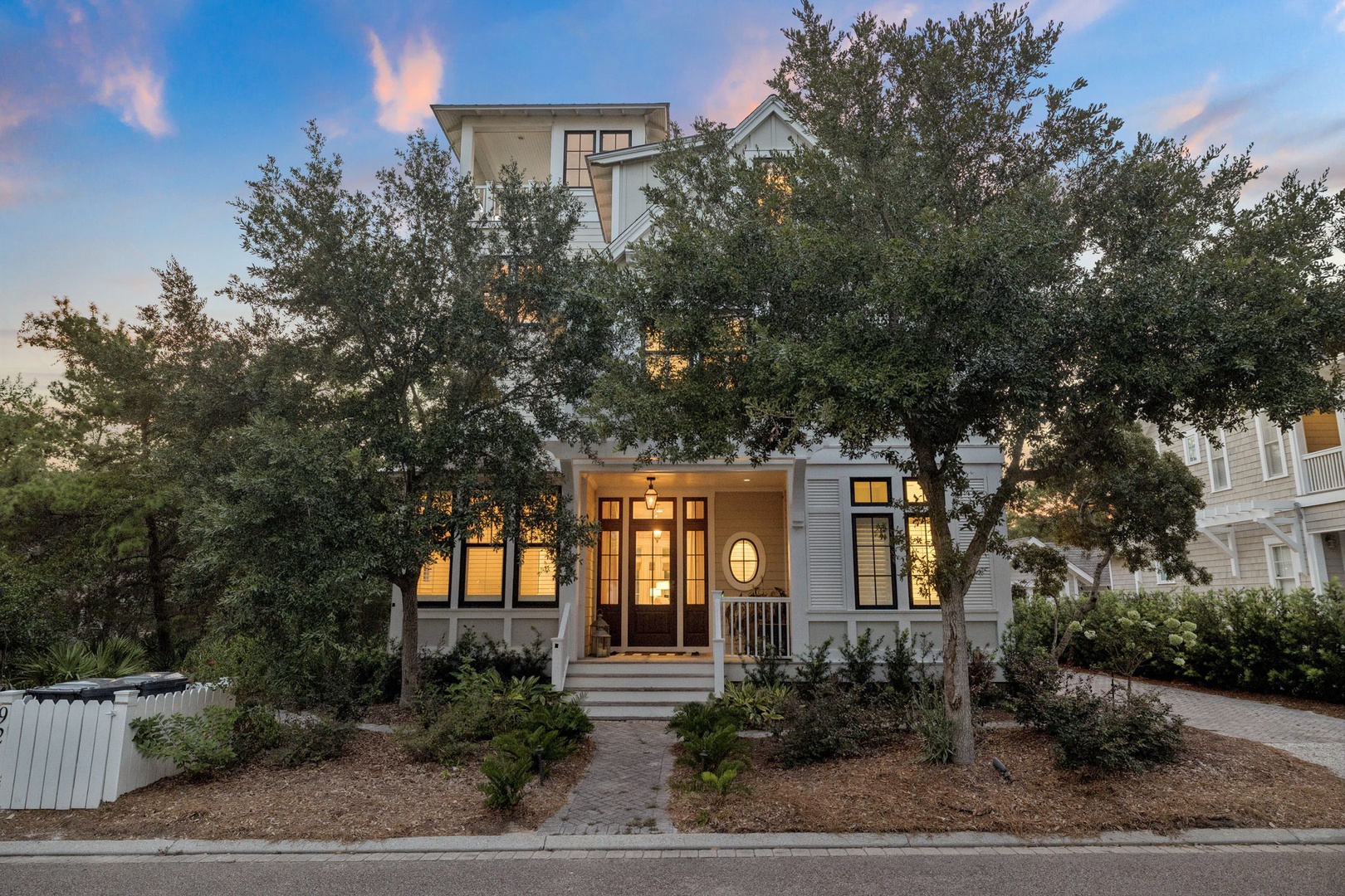A charming coastal home nestled among mature trees, welcoming guests as evening lights glow warmly through its windows in a peaceful residential setting.