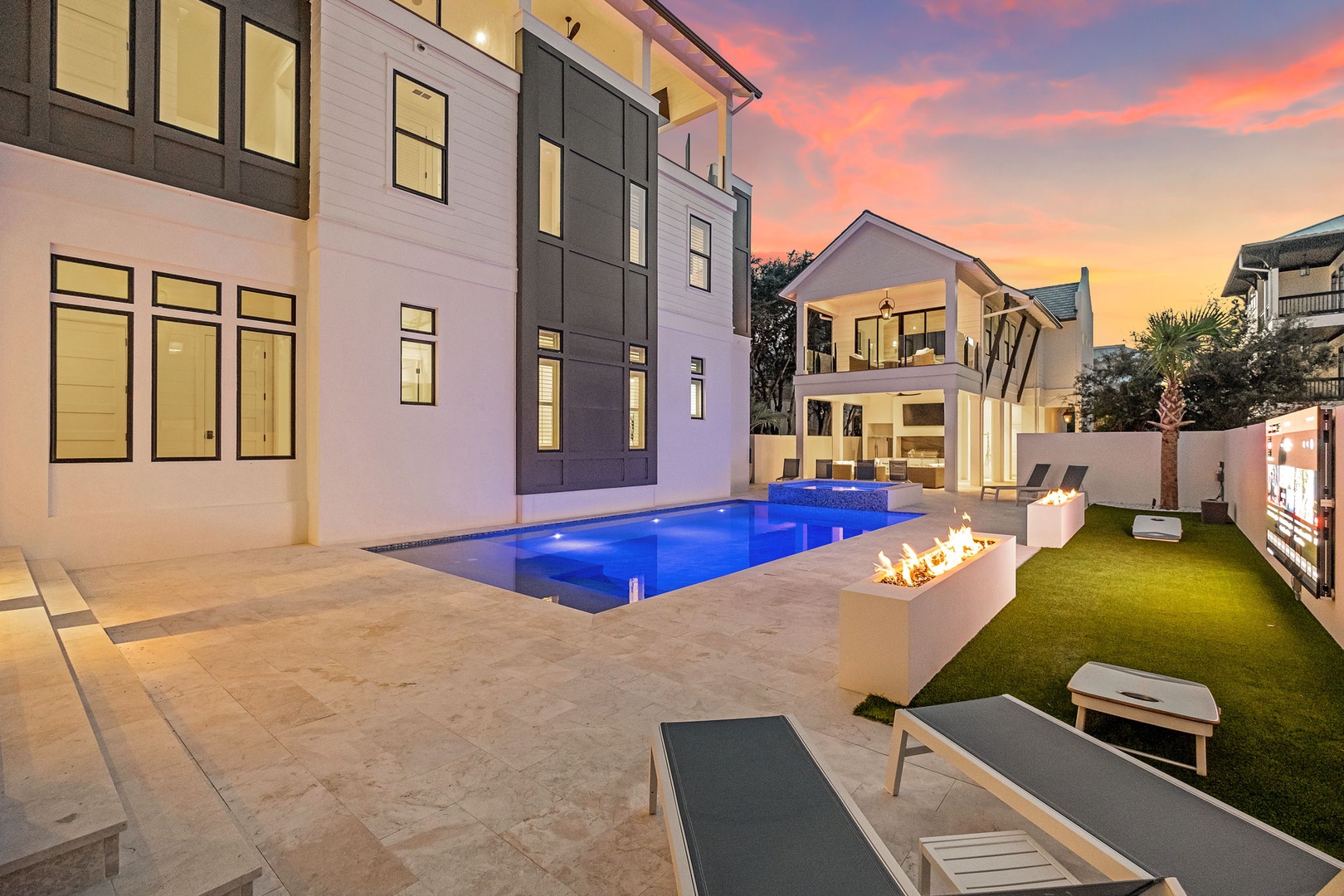 Modern vacation home exterior featuring a stunning illuminated pool and contemporary fire feature beneath a vibrant sunset sky. The sleek architectural design and outdoor lounging areas create an inviting atmosphere for evening relaxation.