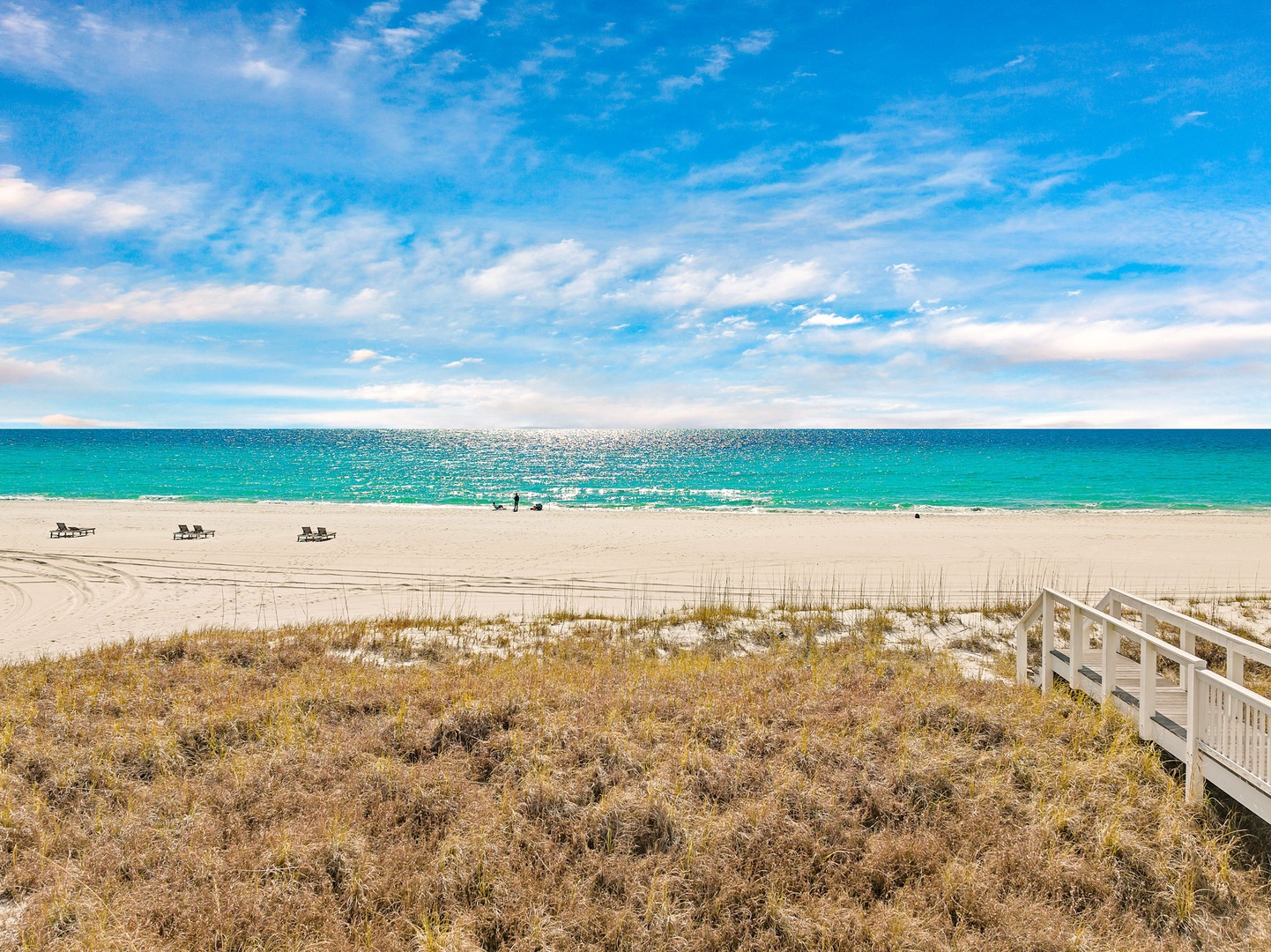 A pristine stretch of white sand beach meets turquoise waters under brilliant blue skies, with lounge chairs ready for your perfect beach day just steps away from the property.