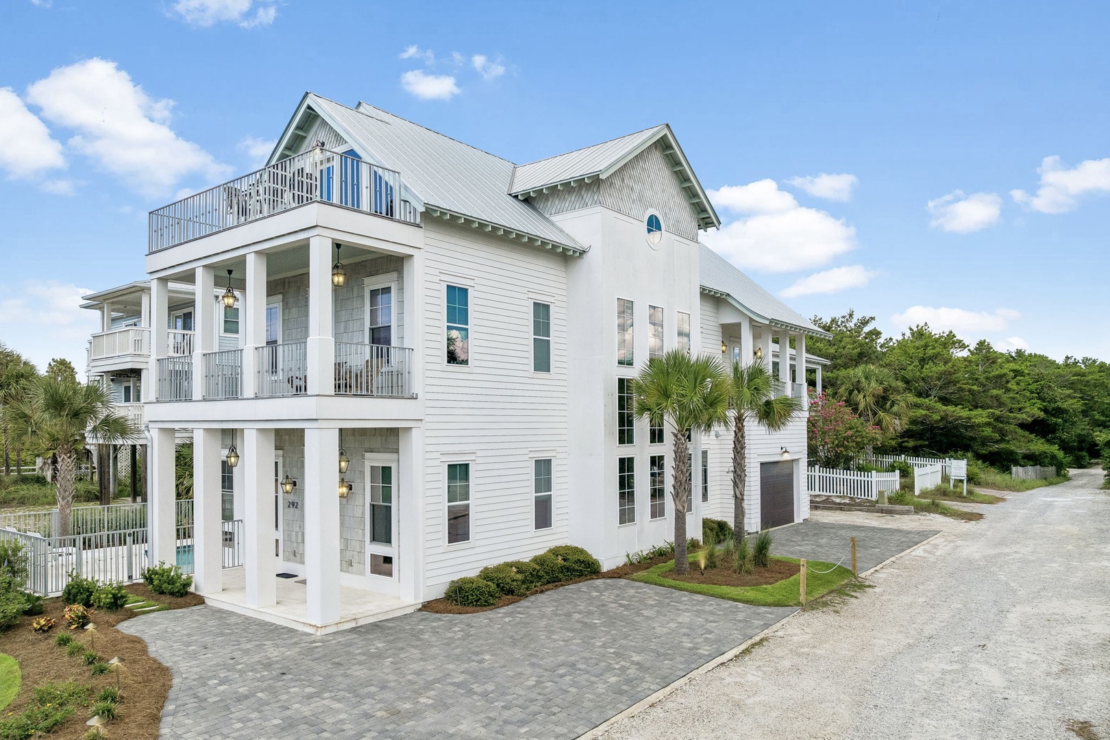 A pristine white coastal-style property with wraparound porches and palm trees, located at the end of a quiet residential street surrounded by lush greenery.