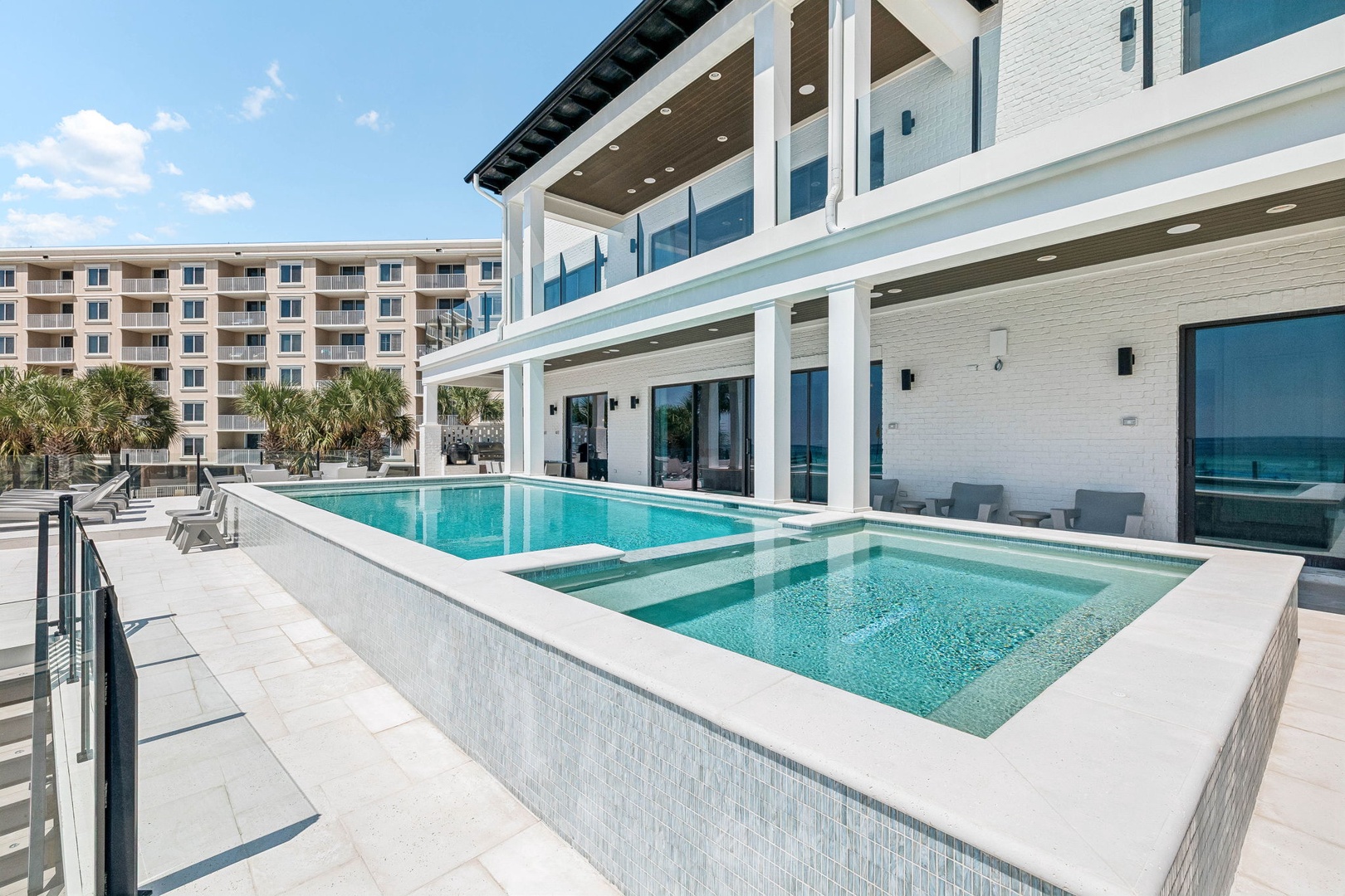 Sophisticated beach home with raised infinity-edge pool and spa overlooking modern multi-level architecture. Sleek white design, glass walls, and tropical palms frame this stunning beachfront property.