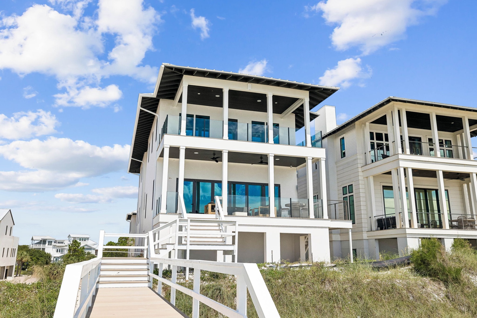 A striking modern beachfront property features expansive windows and wraparound decks across three stories, with direct boardwalk access leading from the sand dunes to your private coastal retreat.