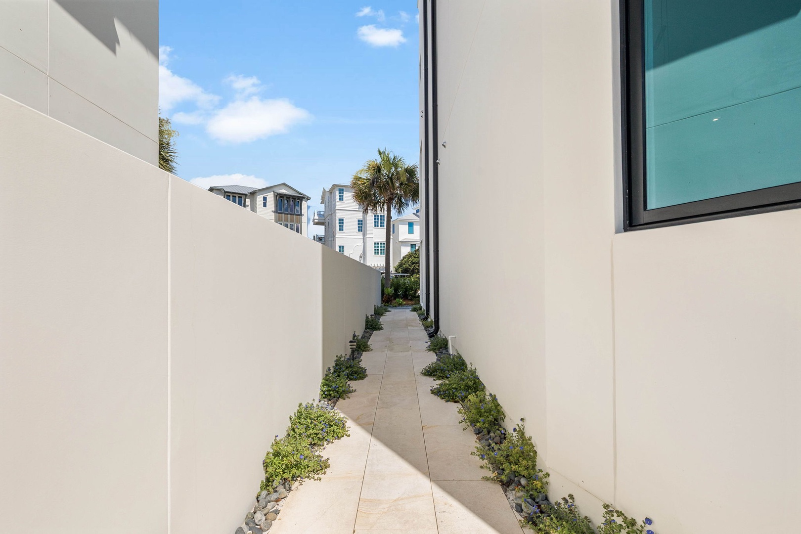 A serene walkway between modern buildings leads you through thoughtfully landscaped greenery, guiding you to your retreat under a clear blue sky with coastal palms nearby.
