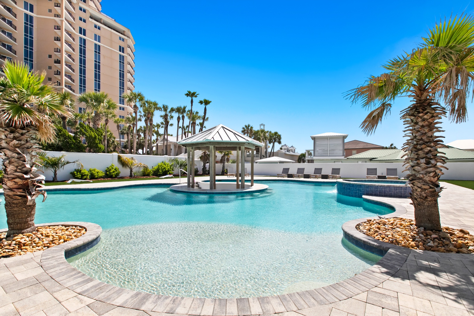 A resort-style pool with crystal-clear water features tropical palm trees, a shaded cabana, and shallow wading areas under bright blue skies—perfect for relaxing in the sun.