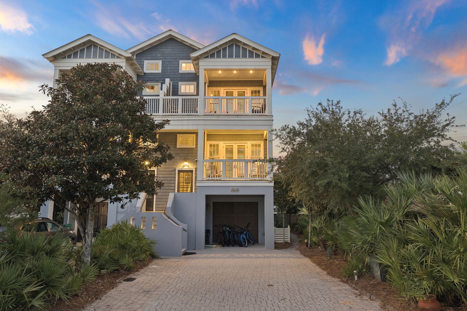 This coastal-style property features classic beach architecture, spacious balconies overlooking the surrounding area, and lush tropical landscaping that frames the grand entrance as the sun sets over the neighborhood.