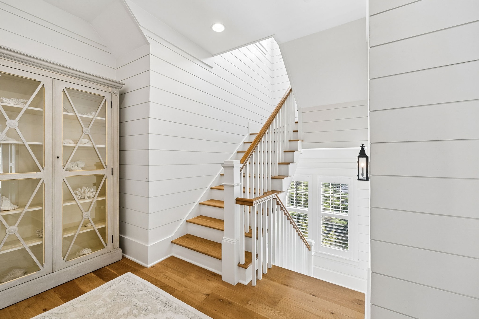 A charming coastal stairway welcomes you with shiplap walls and warm wood tones, blending cottage comfort with timeless elegance. Natural light filters through nearby windows, creating an inviting space to move through your home.