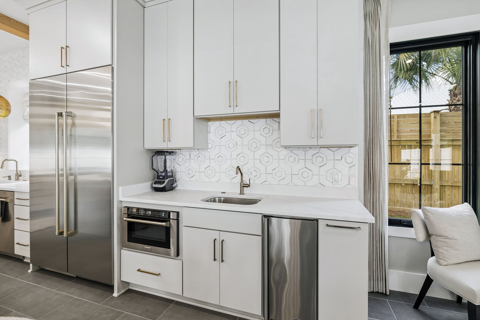 Your modern kitchen is ready for anything you'd like to prepare, with sleek white cabinetry, stainless appliances, and stylish hexagonal tile backsplash. Natural light pours in through large windows overlooking palm trees and a private fenced patio.