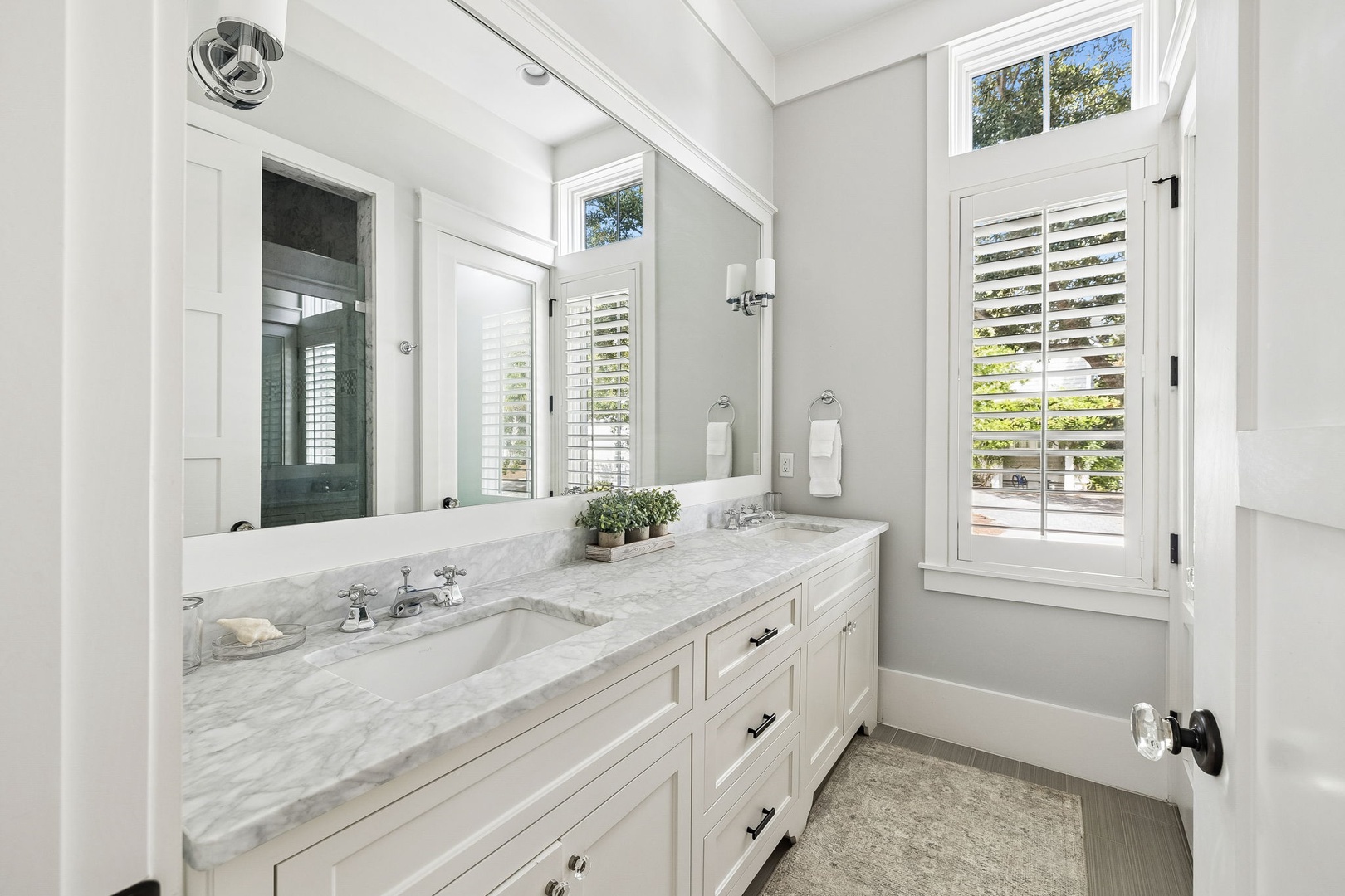 Step into your bright, airy bathroom where natural light floods through plantation shutters, revealing elegant marble countertops and a refreshing garden view. The classic white finishes and thoughtful touches create your perfect morning retreat.