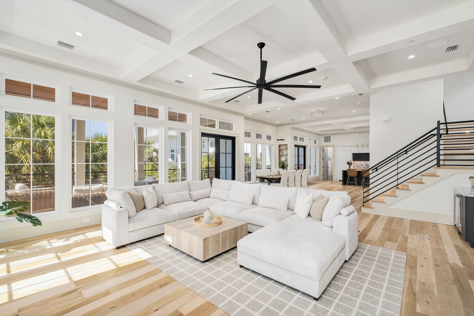 Sink into plush white sectional seating beneath soaring coffered ceilings, where natural light floods your spacious retreat through floor-to-ceiling windows.