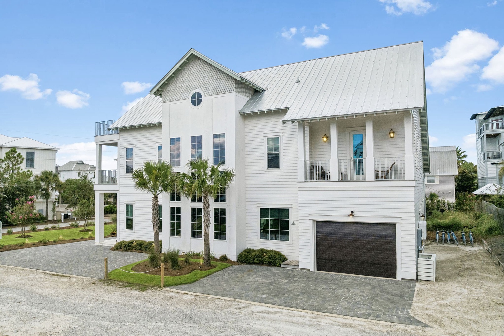 A stunning white coastal property featuring spacious covered porches and tropical palm landscaping, nestled among other beach homes in a peaceful residential area.