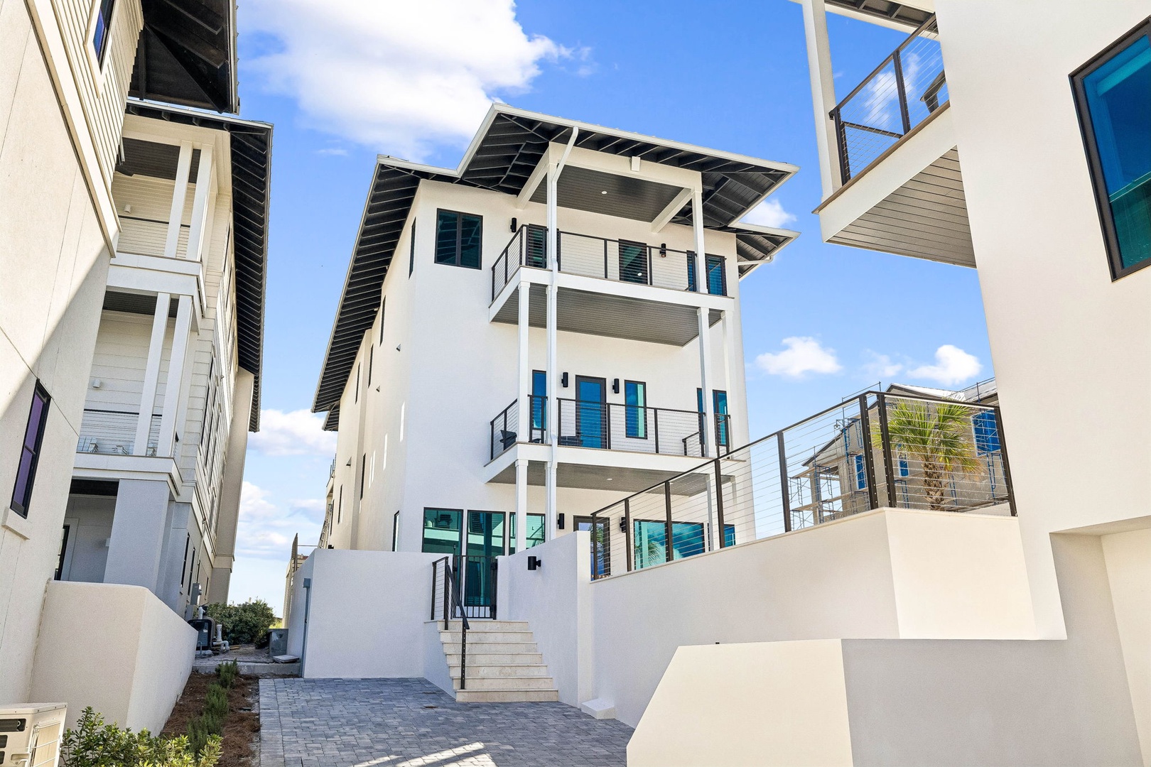 A modern three-story coastal property featuring multiple private balconies and a rooftop deck, showcasing contemporary architecture against a bright blue sky in a residential neighborhood.