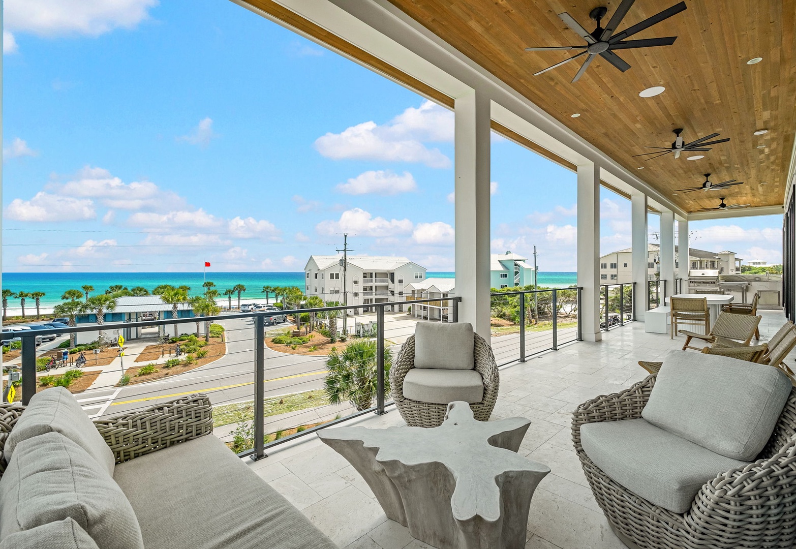 The covered balcony showcases expansive views of turquoise waters and coastal surroundings, framed by modern glass railings and natural wood ceilings with ceiling fans for comfort.