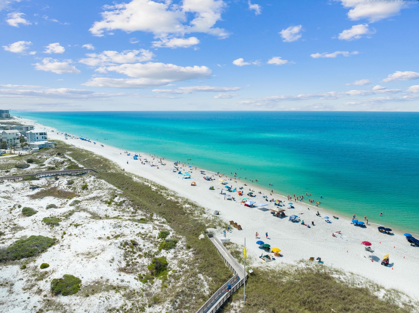 Pristine white sand beach stretches along turquoise waters, with beachgoers enjoying umbrellas and activities on this stunning Gulf Coast shoreline.