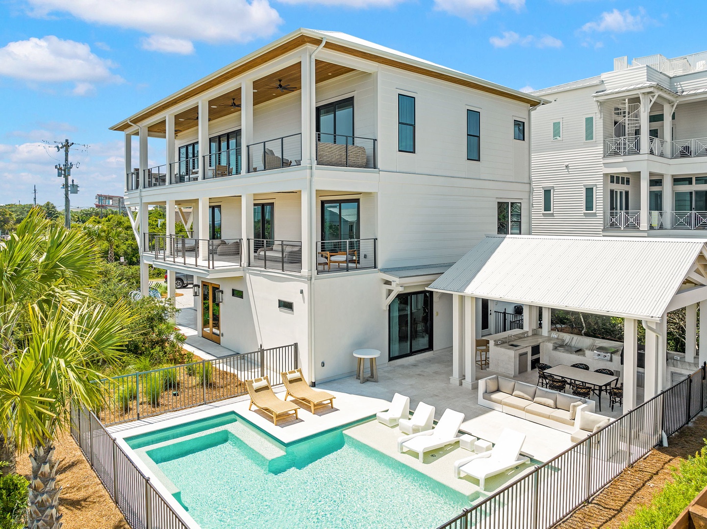 A stunning three-story coastal retreat featuring spacious covered balconies, a private pool with sun loungers, and an outdoor kitchen with dining pavilion. The property sits in a peaceful neighborhood surrounded by lush greenery.