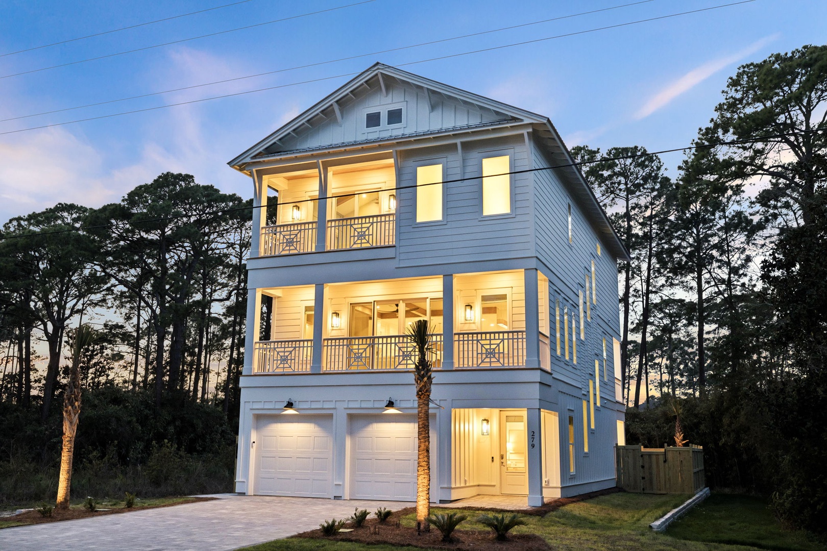 Three-story coastal home with covered balconies and two-car garage, nestled among mature trees in a peaceful wooded setting.