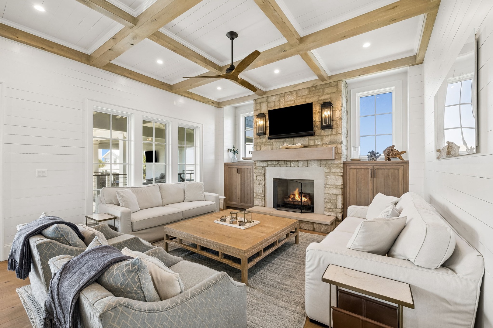 Unwind in your serene living room where exposed beams crown vaulted ceilings and a stone fireplace radiates warmth. Sink into plush seating, gather around the rustic coffee table, and enjoy bright natural light streaming through elegant windows.