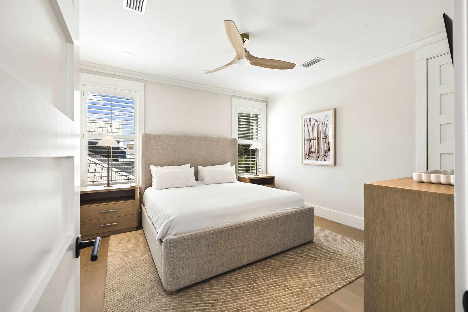 Unwind in this serene bedroom featuring crisp linens, modern ceiling fan, and natural light streaming through plantation shutters. Your coastal retreat awaits with soft textures and calming tones.