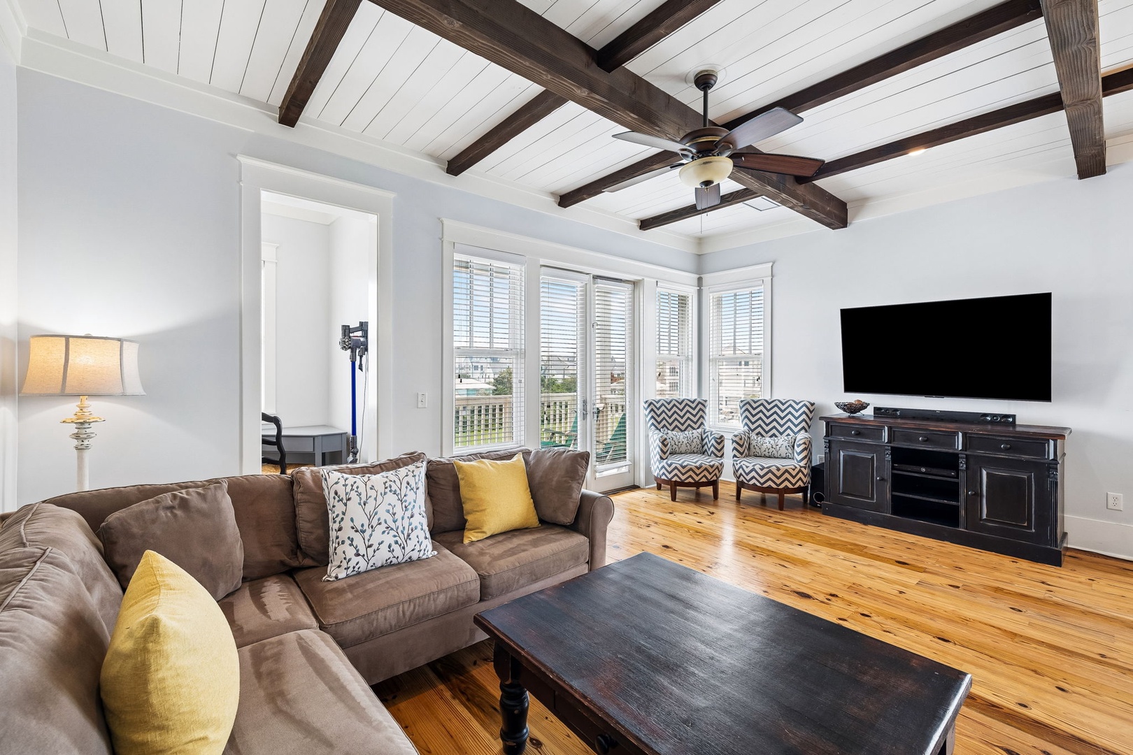 Unwind on plush seating under dramatic beamed ceilings in your spacious living room, where natural light streams through plantation shutters and a ceiling fan keeps you comfortable year-round.