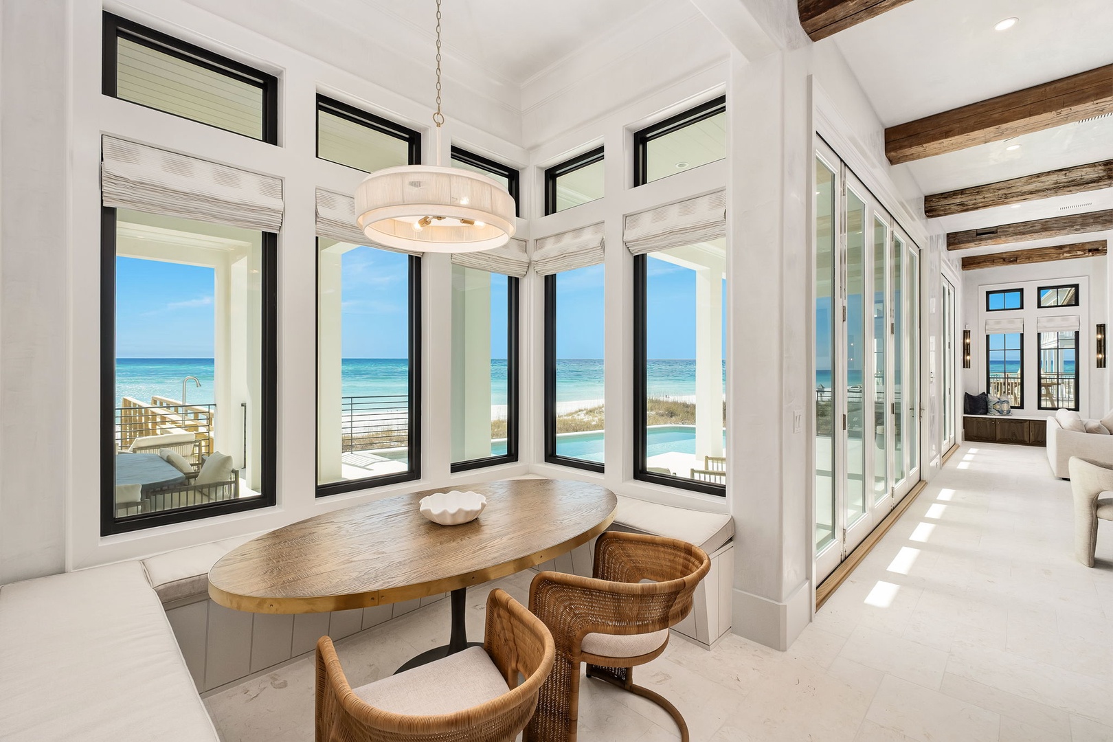 Your morning coffee awaits at this stunning breakfast nook, where floor-to-ceiling windows frame endless ocean views and warm wood beams create coastal elegance.