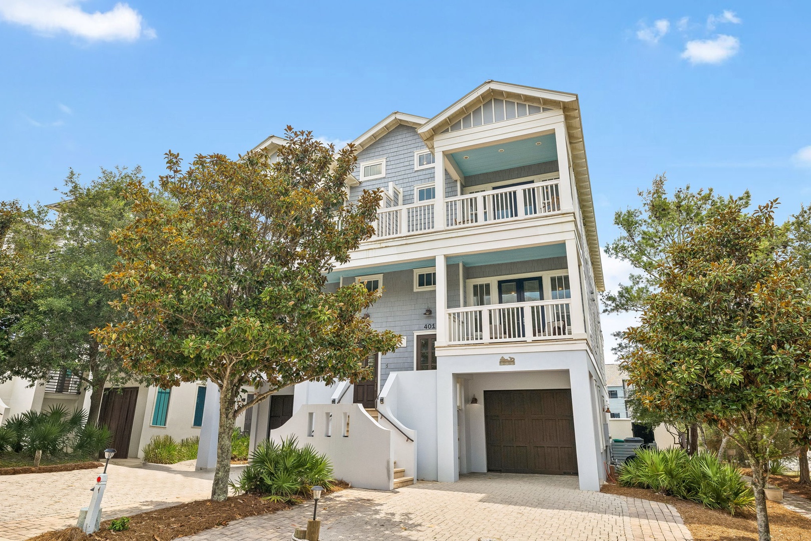 This three-story coastal home features multiple balconies with white railings, nestled among mature trees in a quiet neighborhood setting with paved driveway and ground-level garage.