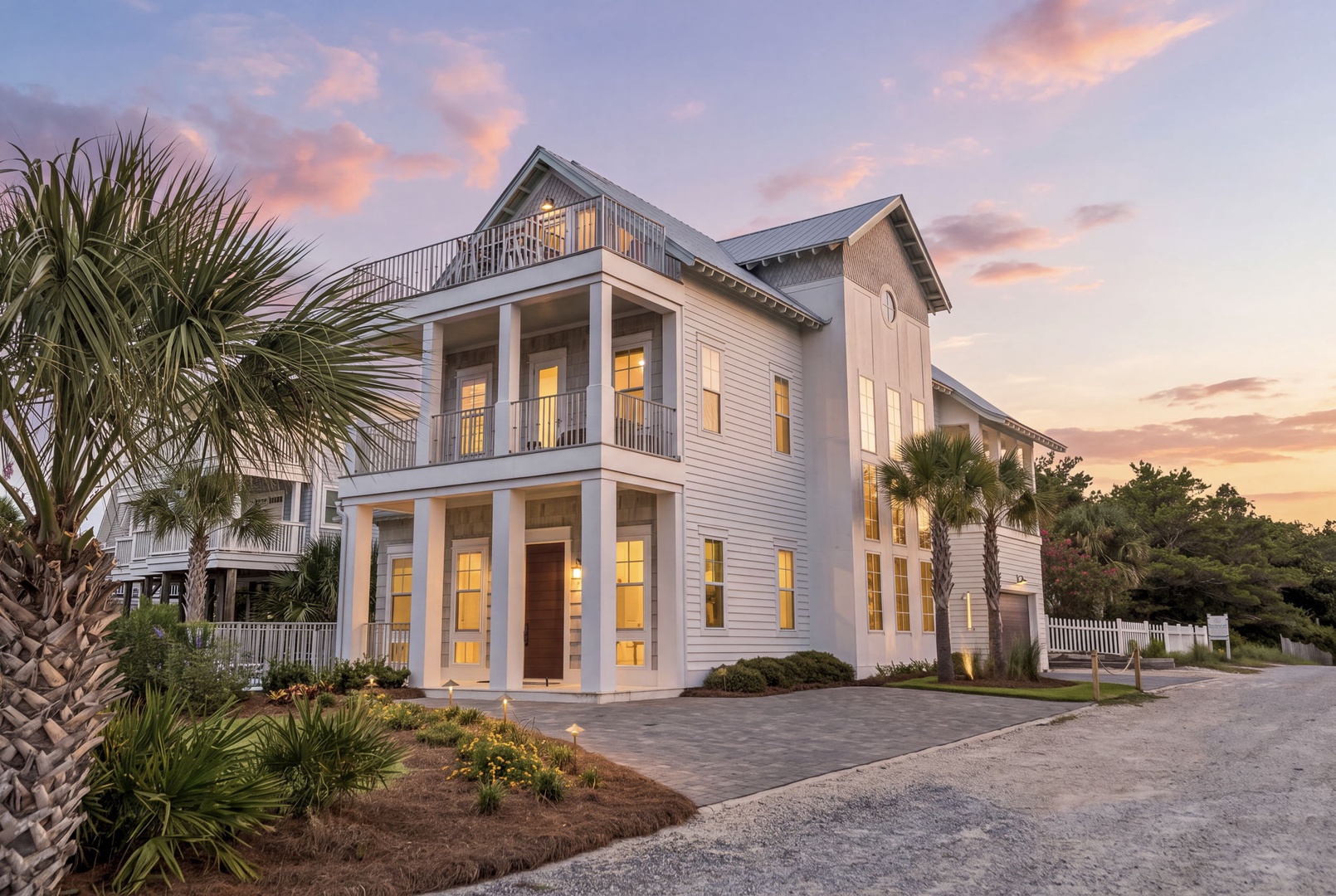 This elegant coastal retreat sits beneath a beautiful sunset sky, surrounded by tropical palms and lush landscaping that welcome guests to their peaceful getaway.