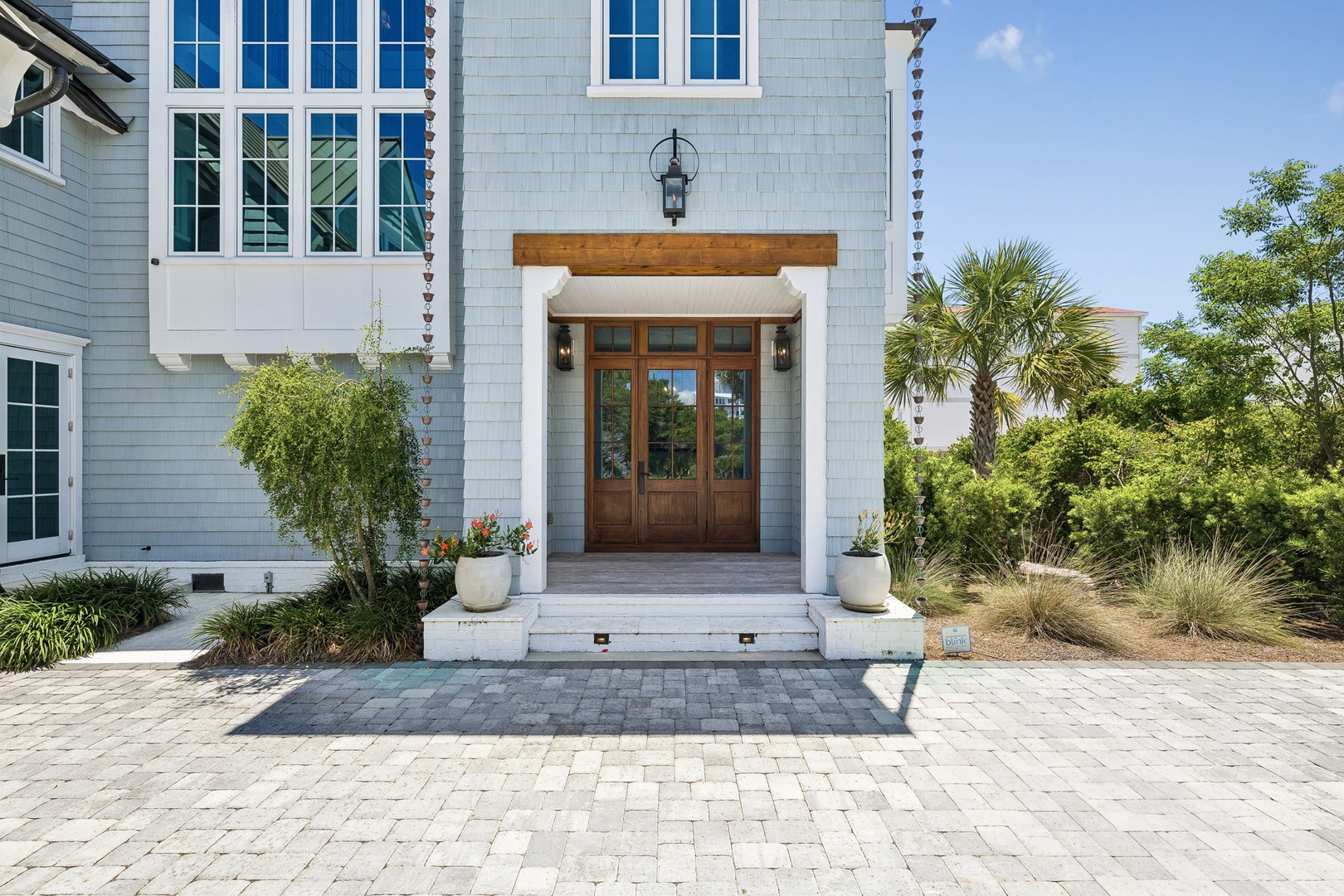 Elegant coastal property entrance featuring light gray shingle siding, warm wood double doors under a natural beam, and manicured landscaping with palm trees and ornamental grasses throughout the surrounding area.