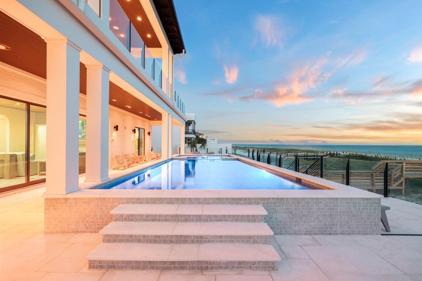 The property's exterior showcases a stunning modern design with expansive glass walls opening to a pristine pool deck, all set against a breathtaking coastal backdrop at twilight.