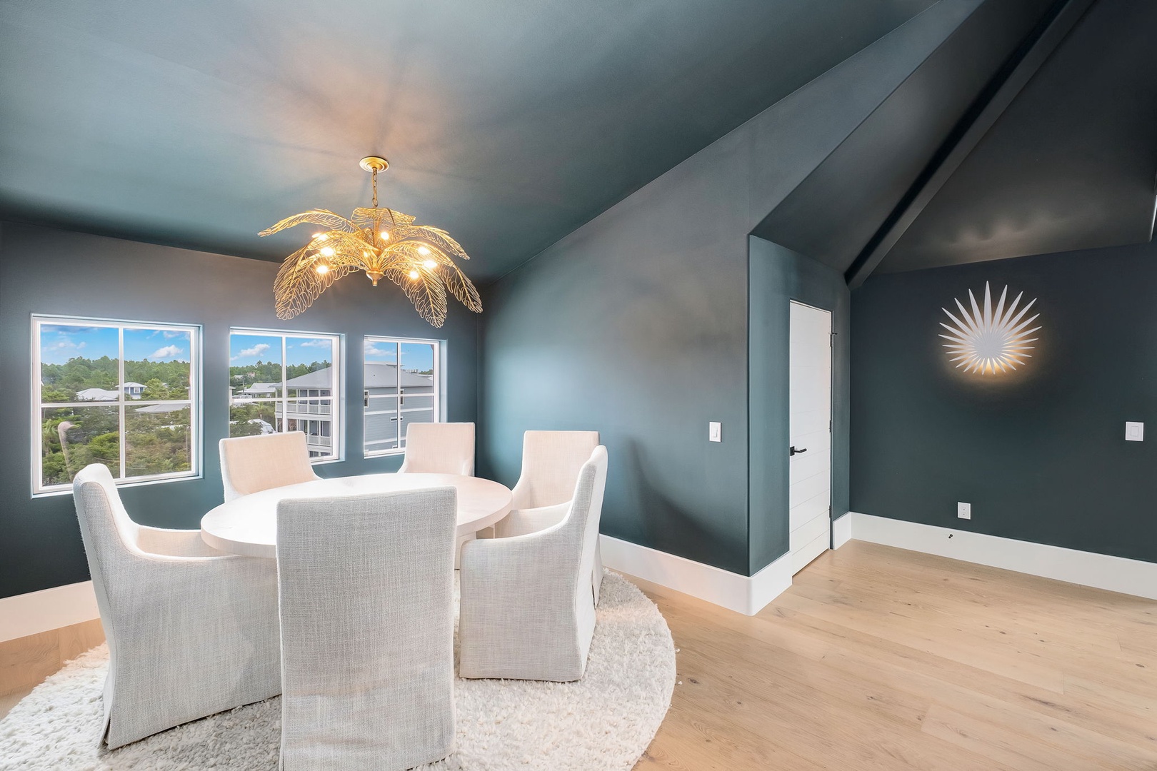 Gather around your elegant dining table beneath a stunning chandelier, where rich teal walls and warm lighting set the perfect mood for memorable meals. Large windows invite natural light to brighten your celebrations.