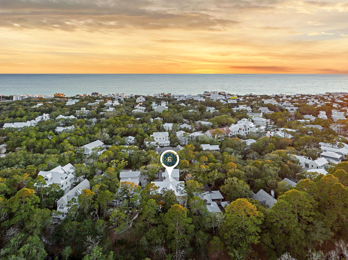 Aerial view of a charming coastal neighborhood nestled among lush trees, with the ocean stretching to the horizon under golden sunset skies.