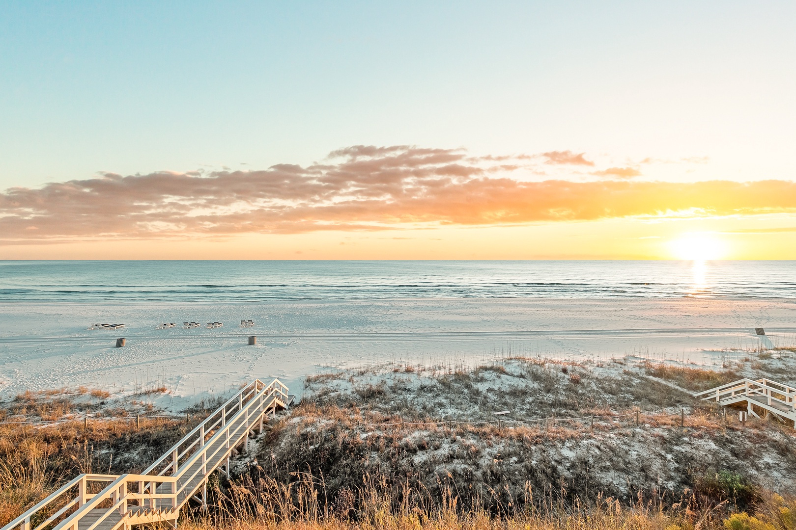Golden sunrise paints the sky above pristine beach access with wooden boardwalk leading to sandy shores and gentle waves.