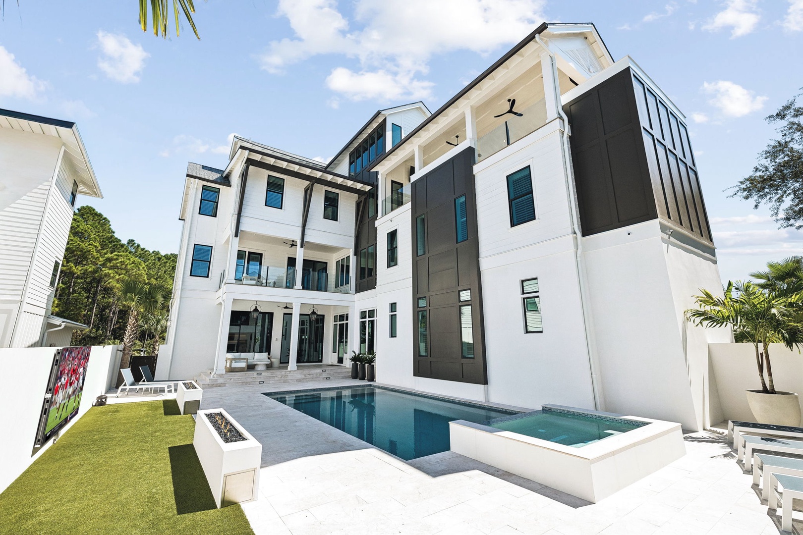 A striking modern property showcasing sleek contemporary architecture, with a sparkling pool and elevated hot tub surrounded by tropical landscaping under a bright, sunny sky.