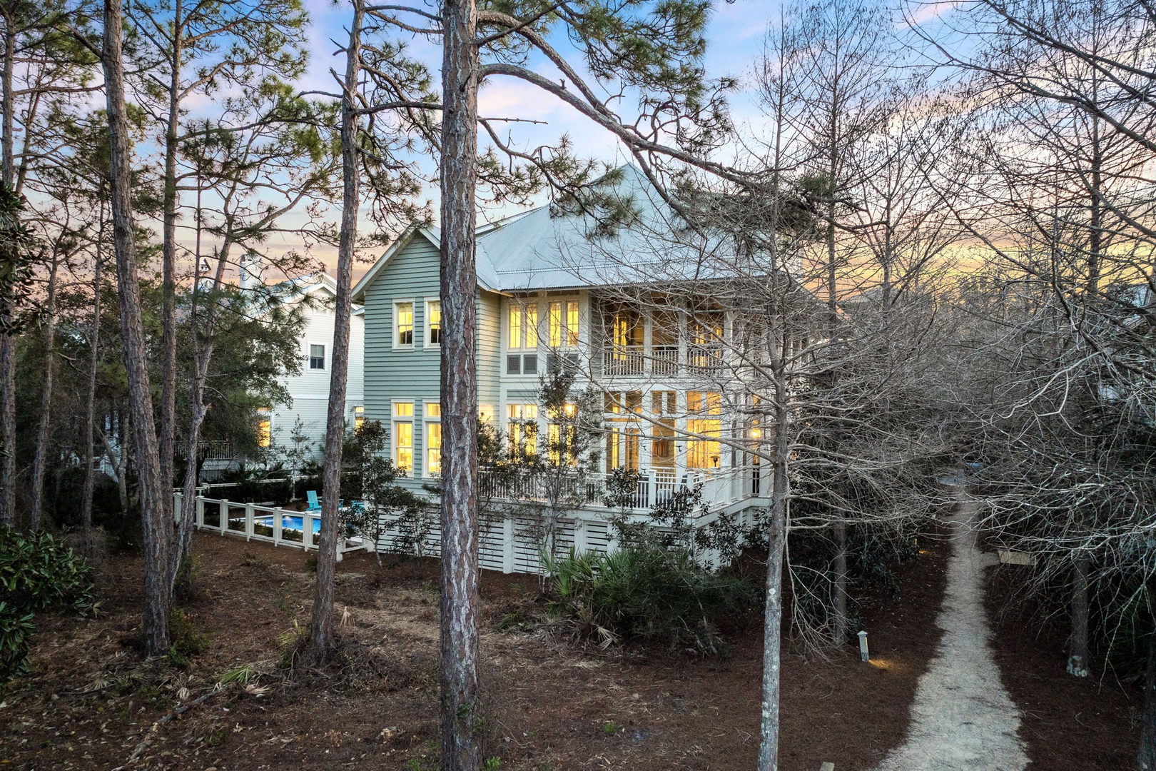 A charming coastal retreat nestled among towering pines, where warm lights beckon through large windows and a welcoming wraparound porch awaits.