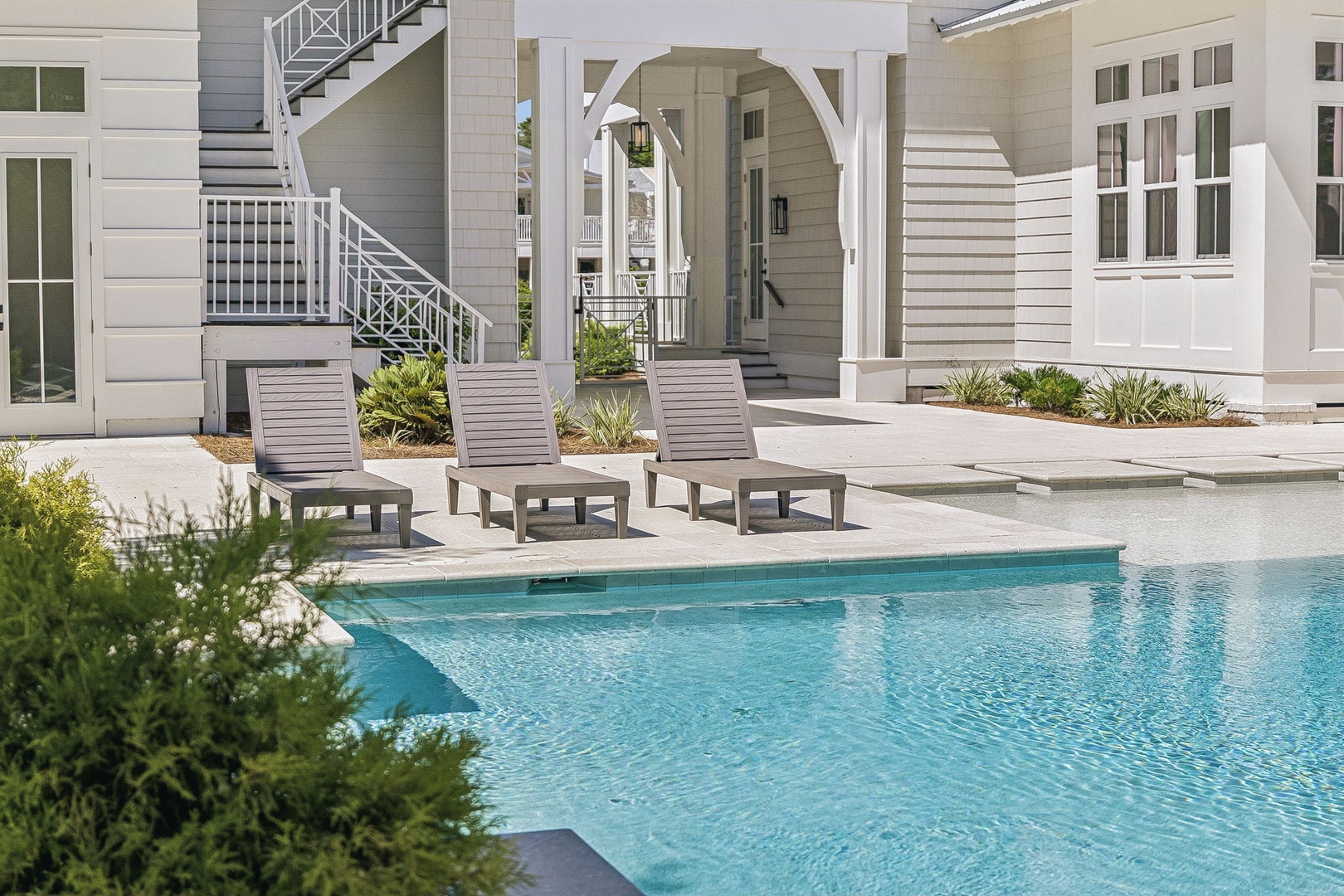 Three poolside loungers invite you to unwind in your private oasis, surrounded by elegant coastal architecture and lush greenery. The sparkling water and sun-drenched deck set the stage for blissful relaxation.