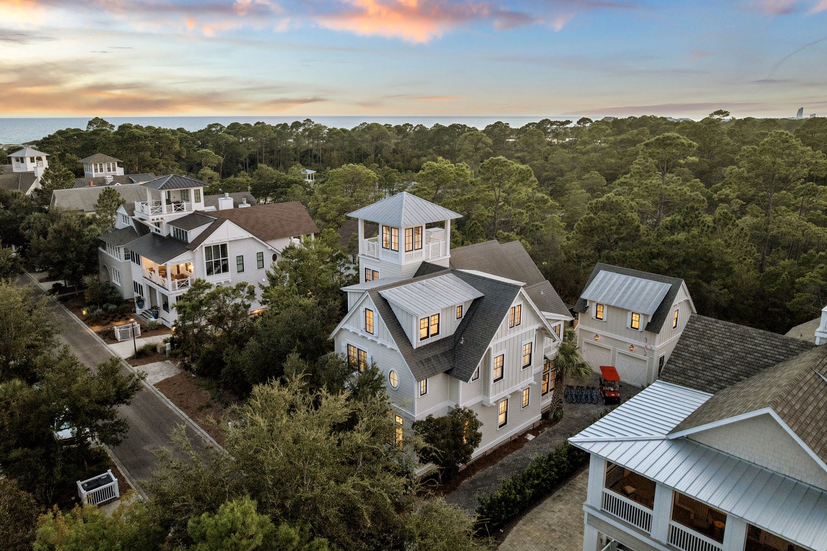 An aerial view captures the property nestled within a lush coastal community at sunset, surrounded by mature trees and neighboring homes.