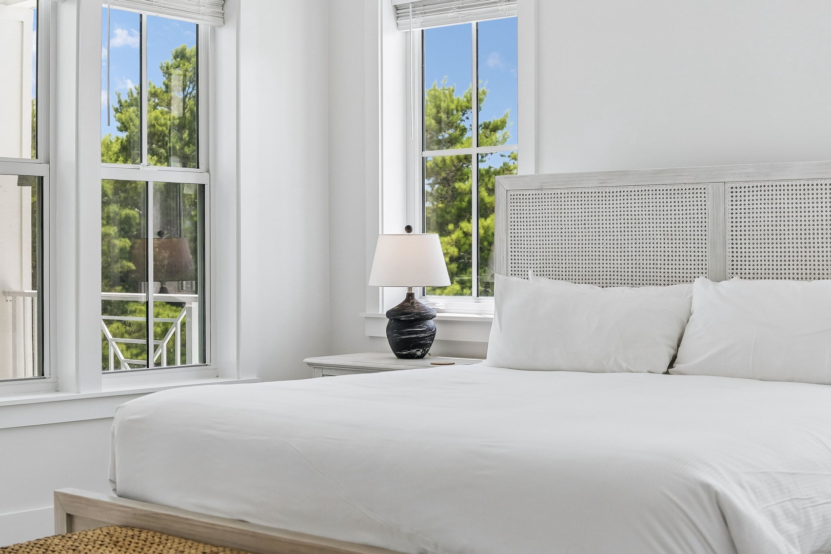 Light floods your serene bedroom through tall windows framing lush greenery. Sink into crisp white linens and breathe in the peaceful coastal atmosphere that awaits you each morning.