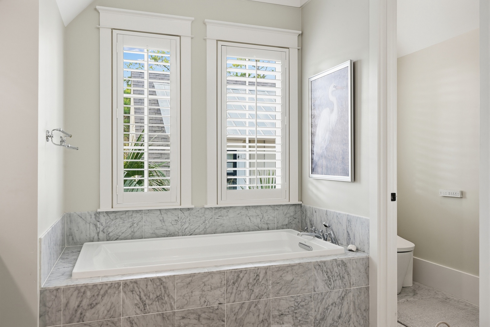Sink into your own private sanctuary—marble-wrapped soaking tub beneath elegant shuttered windows, where warm light filters through for your ultimate relaxation.