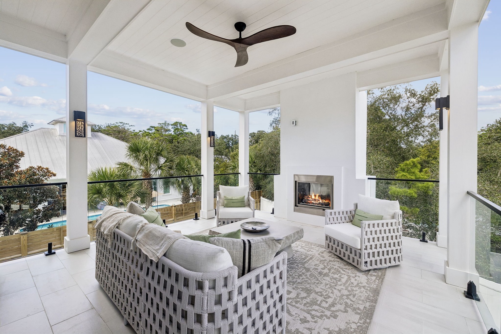 Relax in your covered outdoor lounge with a cozy fireplace, stylish seating, and refreshing views of lush greenery and the sparkling pool just beyond.